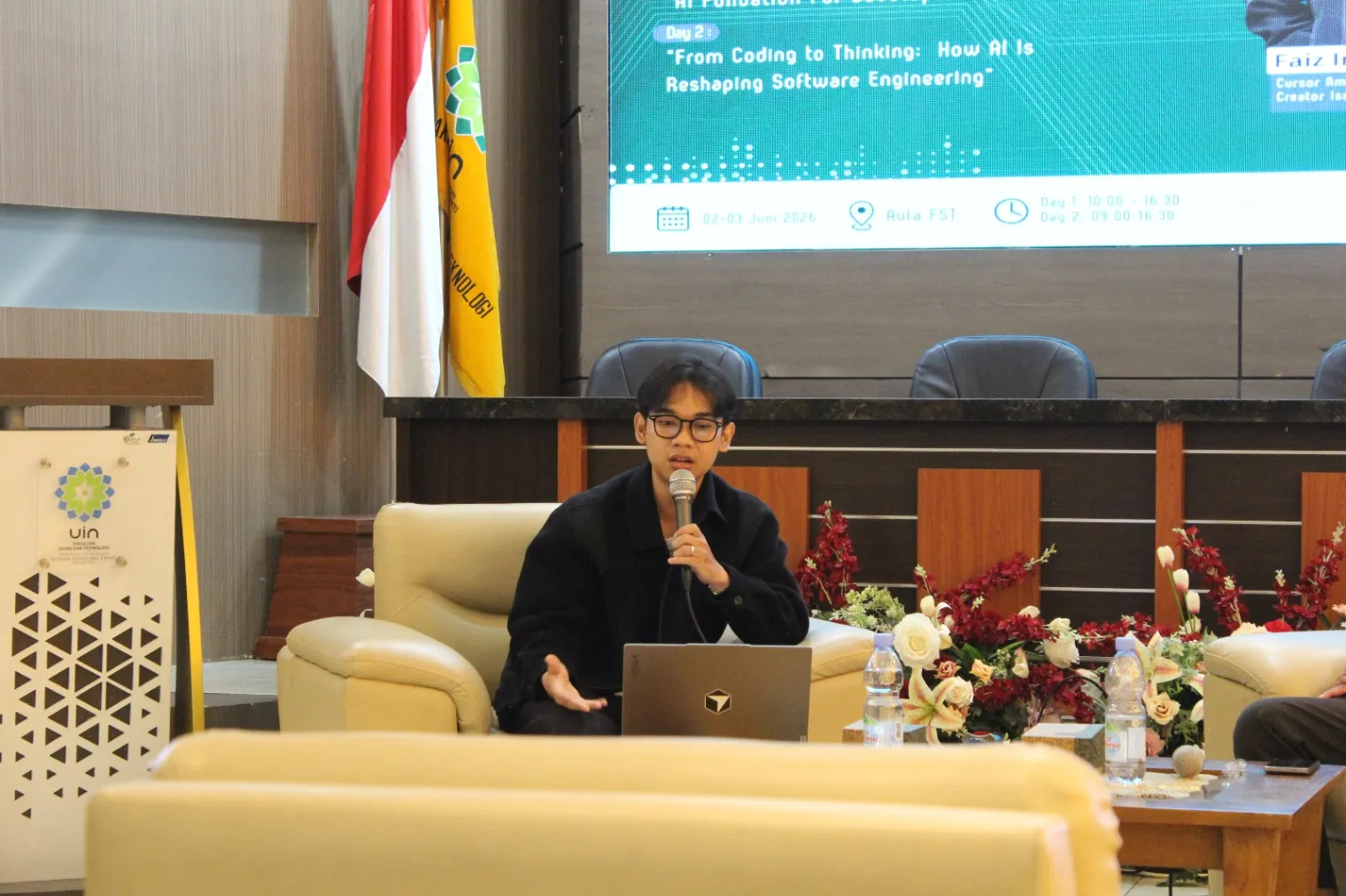 Faiz speaks with a microphone at Aula FST, in front of a screen with the day 2 session title